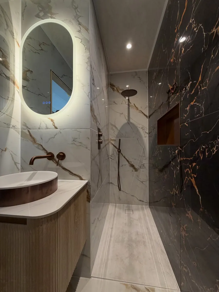Copper vessel basin under an oval backlit mirror, with the shower beyond